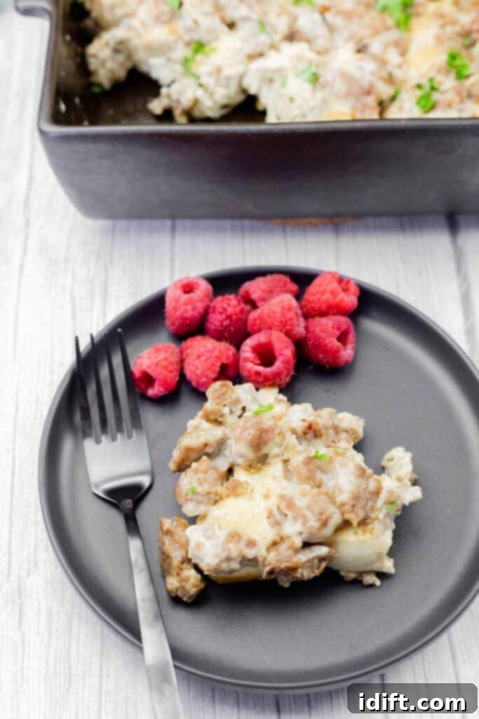 A black plate with a serving of sausage casserole and fresh raspberries, alongside a black fork. A casserole dish is partially visible in the background.