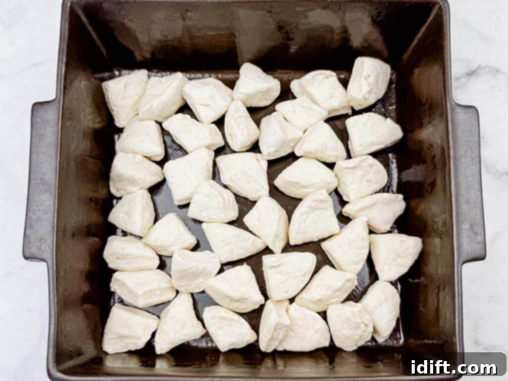 A square baking pan filled with evenly spaced, uncooked biscuit dough pieces, arranged on a white surface.