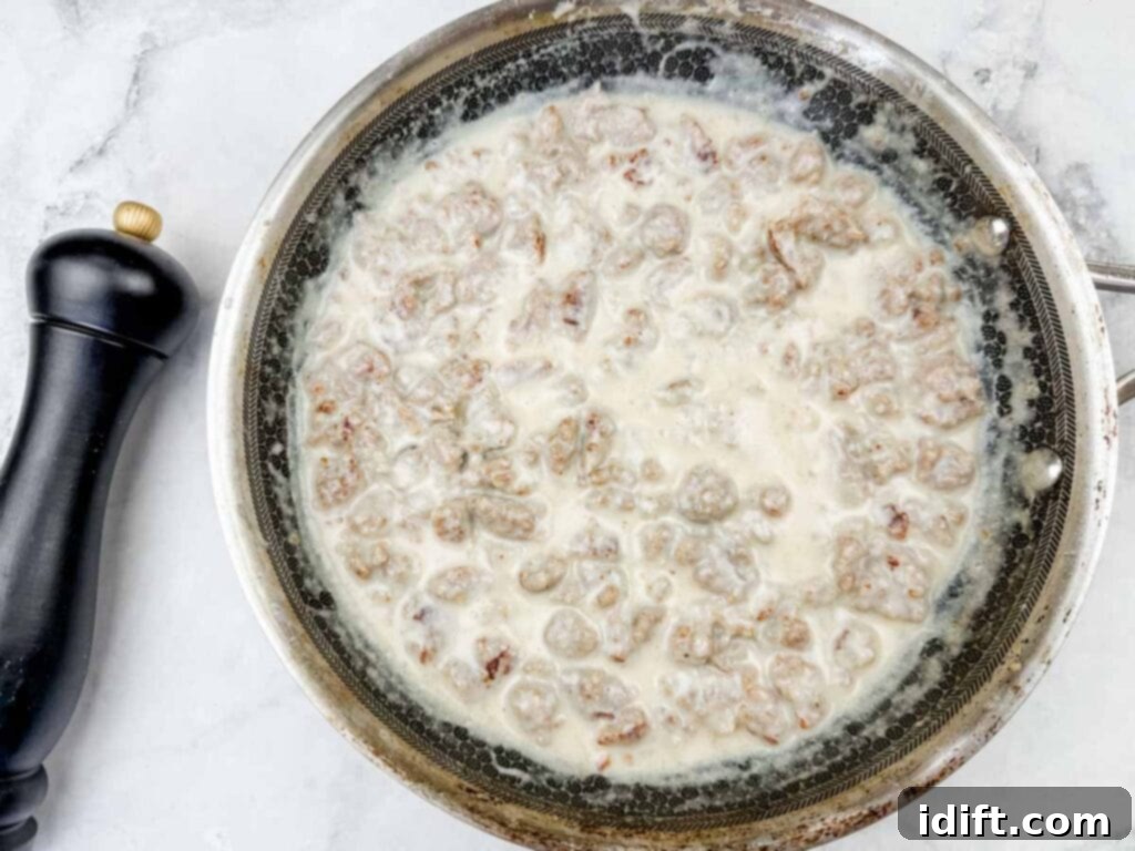 A skillet filled with cooked sausage in creamy white gravy, placed on a marble countertop with a black pepper grinder nearby.
