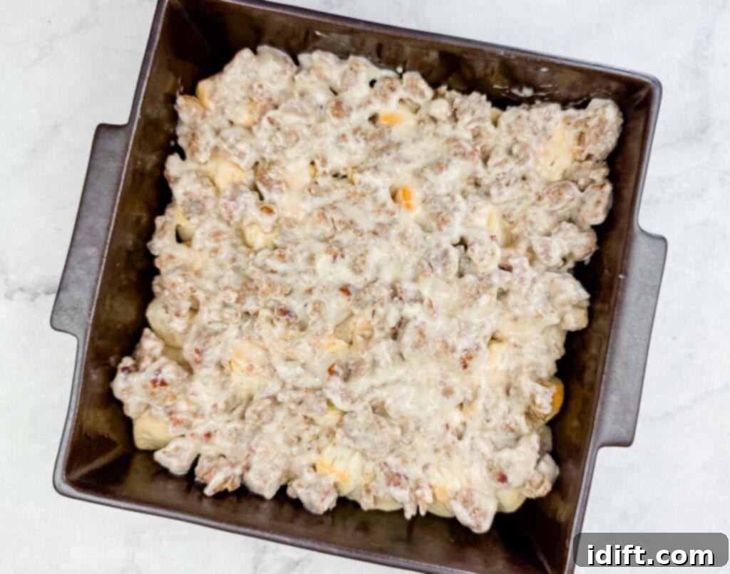 A square baking pan filled with a baked casserole topped with melted cheese and pieces of sausage on a light surface.