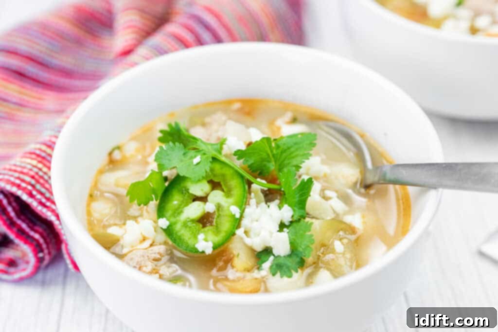 A vibrant bowl of Green Pork Posole Soup garnished with fresh cilantro, thinly sliced jalapeños, and crumbled queso fresco, with a spoon resting in the bowl and a striped kitchen towel in the background, suggesting a cozy meal.