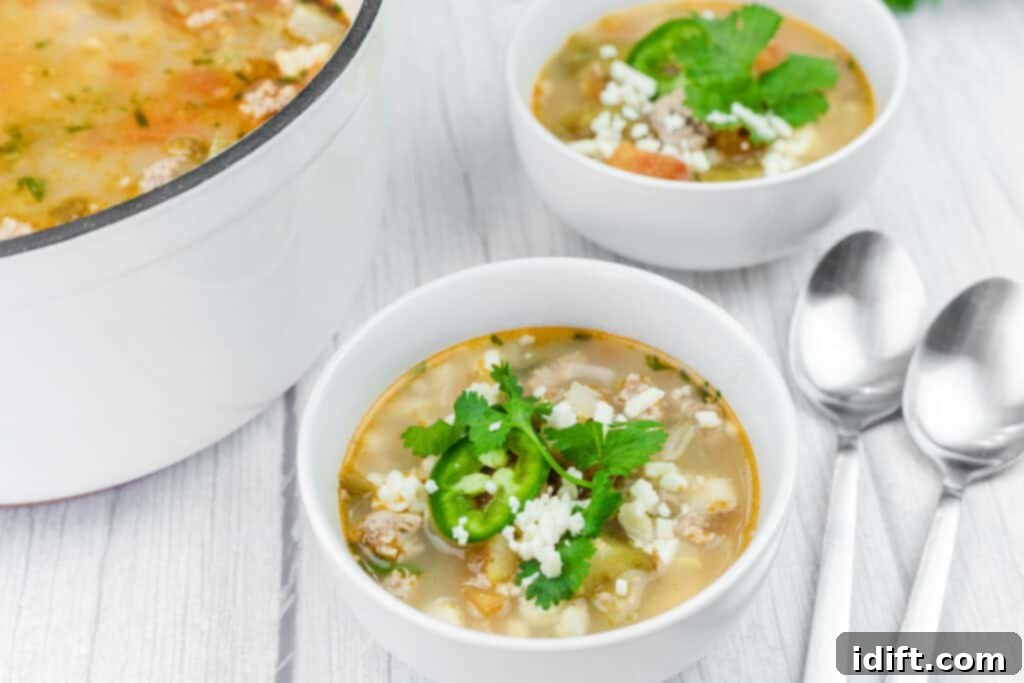 Three perfectly garnished bowls of Green Pork Posole, topped with vibrant cilantro, thinly sliced jalapeños, and crumbled cheese, arranged on a light wooden surface. Two spoons rest beside the bowls, with a large pot of soup partially visible in the background, inviting everyone to enjoy.