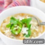 A bowl of Green Pork Posole topped with sliced jalapeño, cilantro, and crumbled cheese, with a spoon in the bowl and a striped cloth in the background.