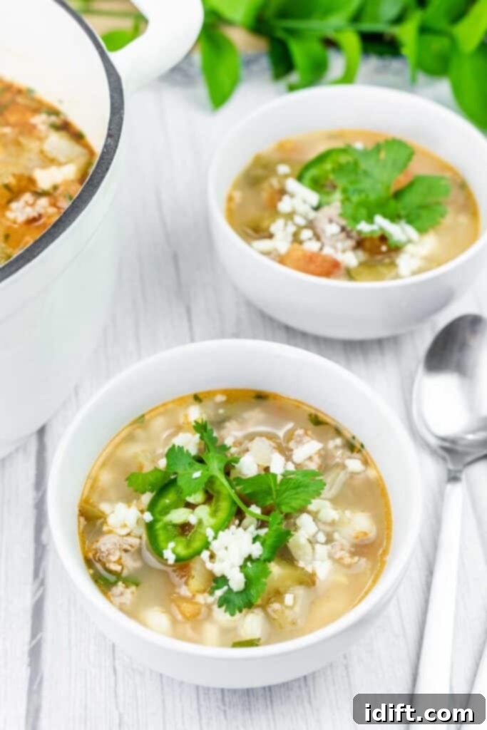 Two generous bowls of Green Pork Posole, richly garnished with fresh cilantro, bright green jalapeño slices, and crumbly white cheese, sit on a rustic wooden table next to a large white pot filled with more soup, ready to be served.