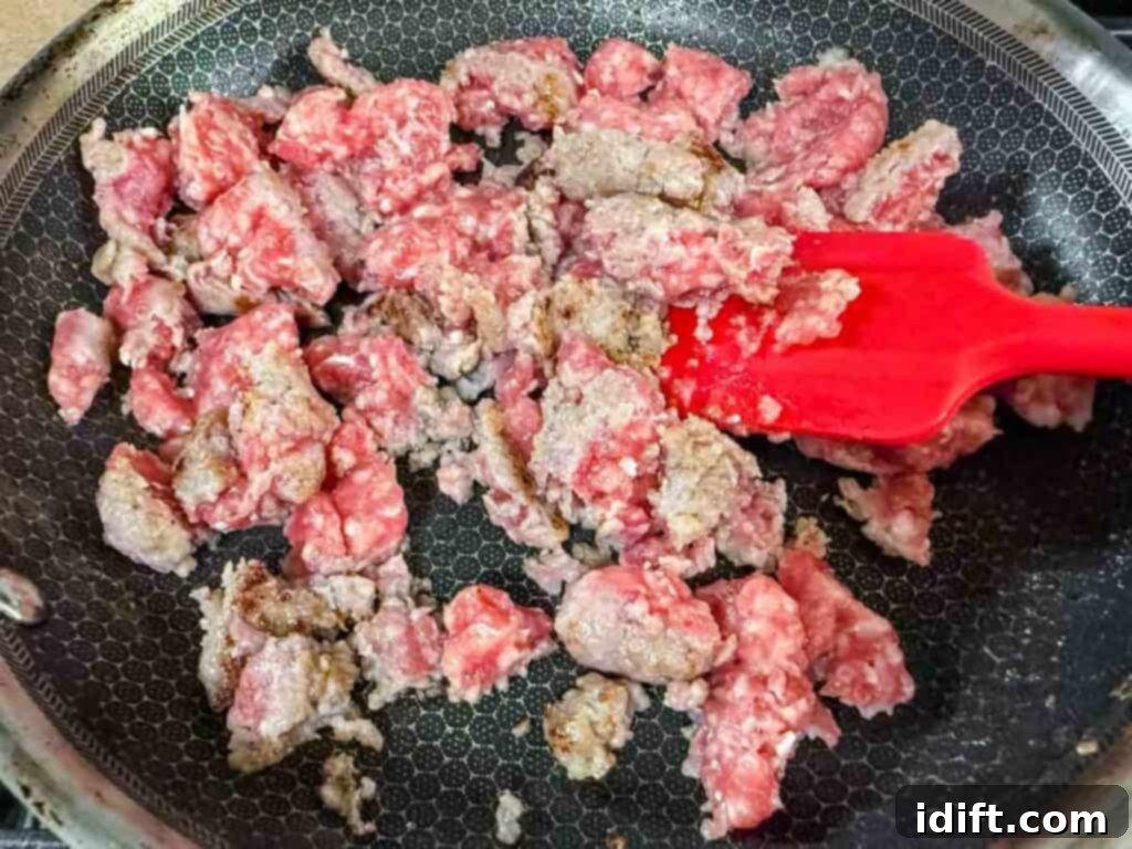 Lean ground pork being expertly browned in a large, deep braiser pan, with a red spatula actively stirring and breaking up the meat into fine crumbles alongside diced onions and bell peppers, setting the savory foundation for the soup.