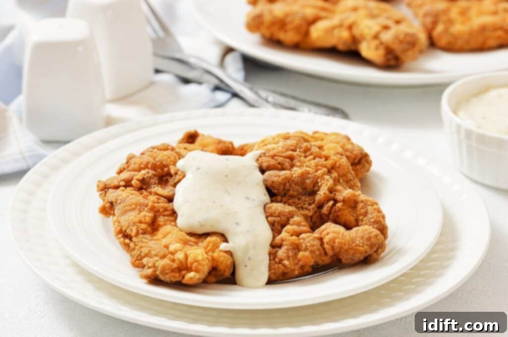 A plate of chicken fried steak topped with white gravy, with another plate and utensils in the background.