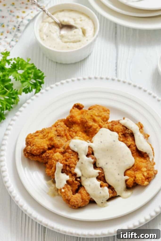 A plate of fried chicken topped with white gravy, served on a white plate with a bowl of gravy and parsley garnish nearby.