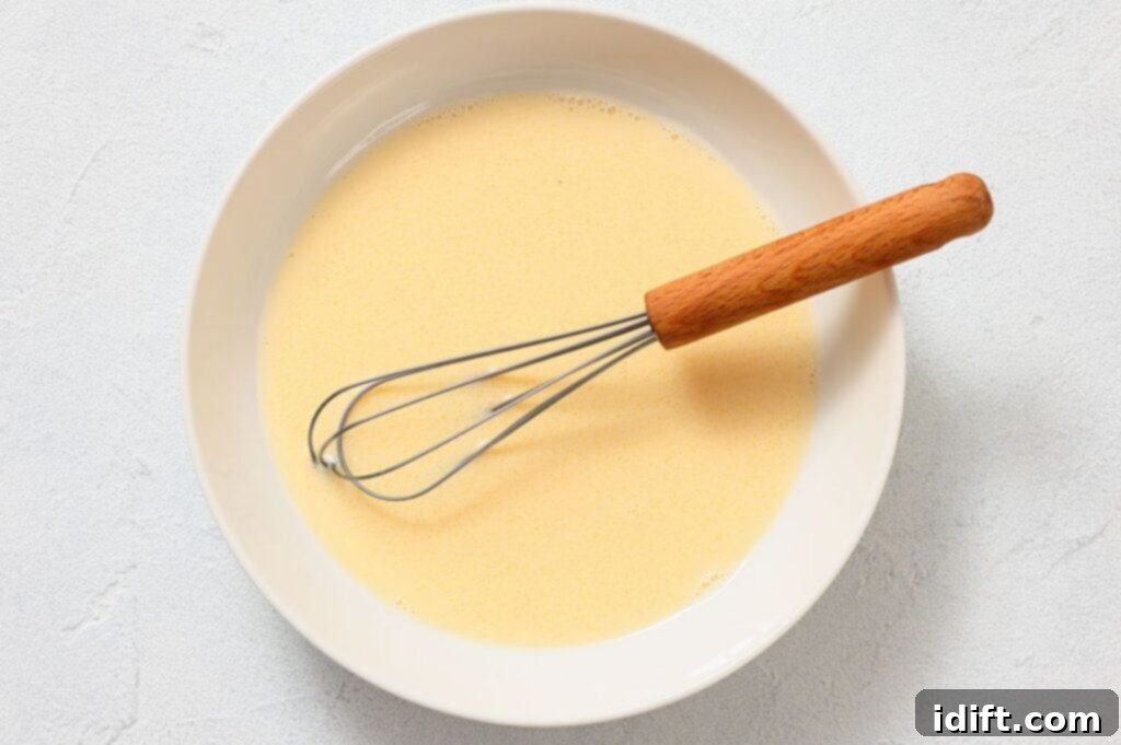 A metal whisk with a wooden handle rests in a white bowl containing a pale yellow liquid mixture on a light surface.