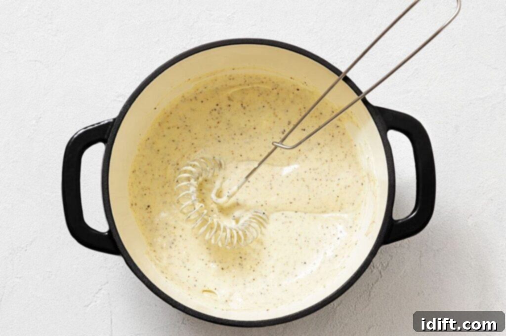A metal whisk rests in a black pot filled with a creamy, seasoned sauce on a light-colored surface.