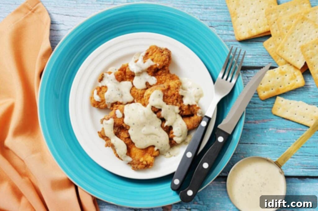 A plate of fried chicken strips topped with white gravy, next to a knife, fork, crackers, a small bowl of gravy, and an orange napkin on a blue wooden surface.
