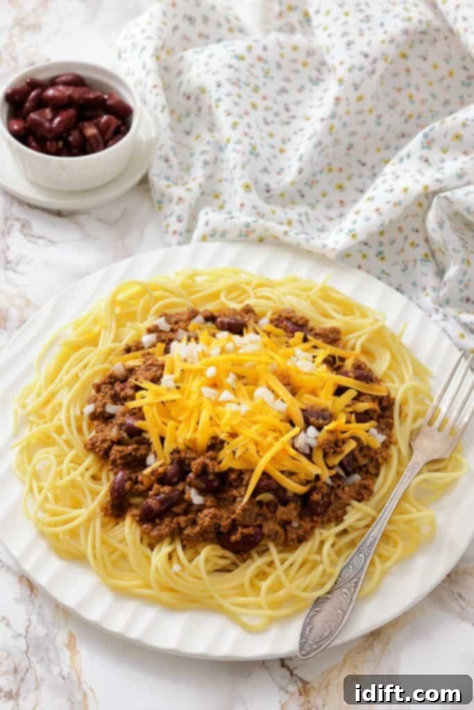The Queen City's Signature Bowl 3 A plate of spaghetti topped with chili, shredded cheddar cheese, and diced onions, with a fork on the side and a bowl of kidney beans in the background.
