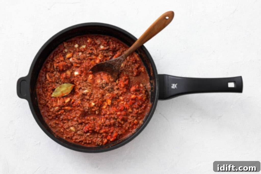 The Queen City's Signature Bowl 6 A black saucepan filled with red meat sauce, diced vegetables, and a bay leaf, with a wooden spoon resting inside, on a white surface.