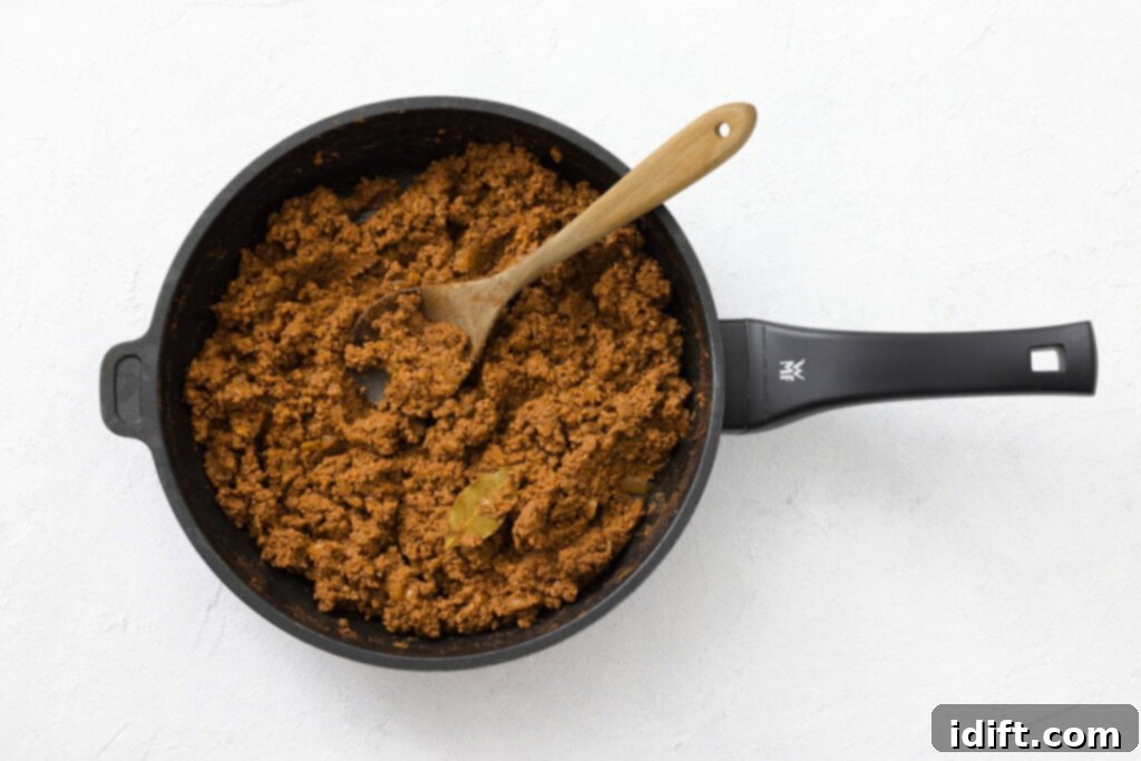 The Queen City's Signature Bowl 7 Cooked ground meat mixture with a bay leaf in a black frying pan, stirred with a wooden spoon, on a white background.
