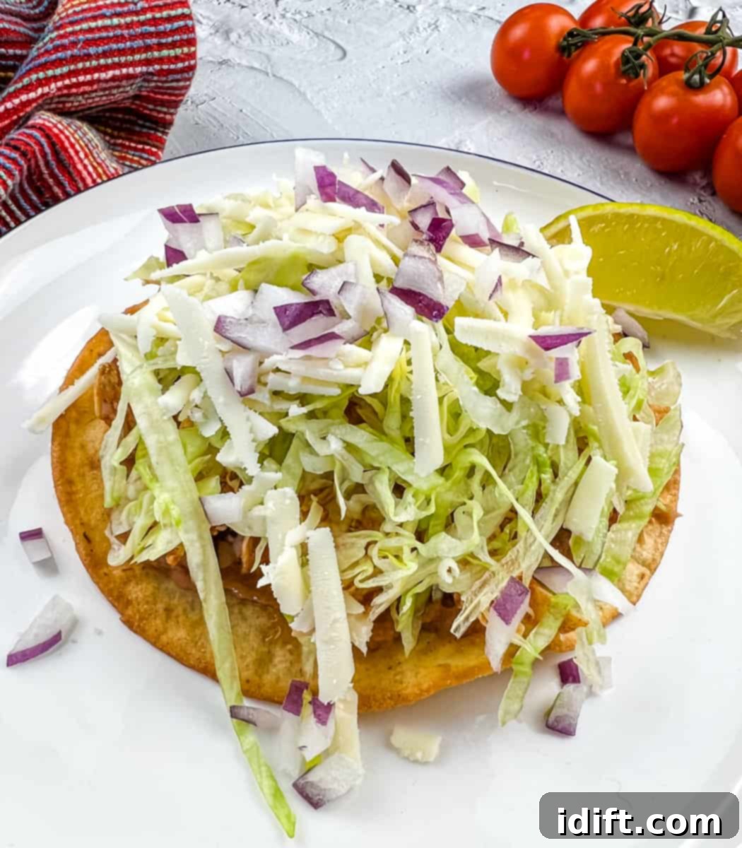 Flavor-packed Chicken Tinga Tostadas on a white plate with fresh tomatoes in the background, showcasing a perfect weeknight meal.