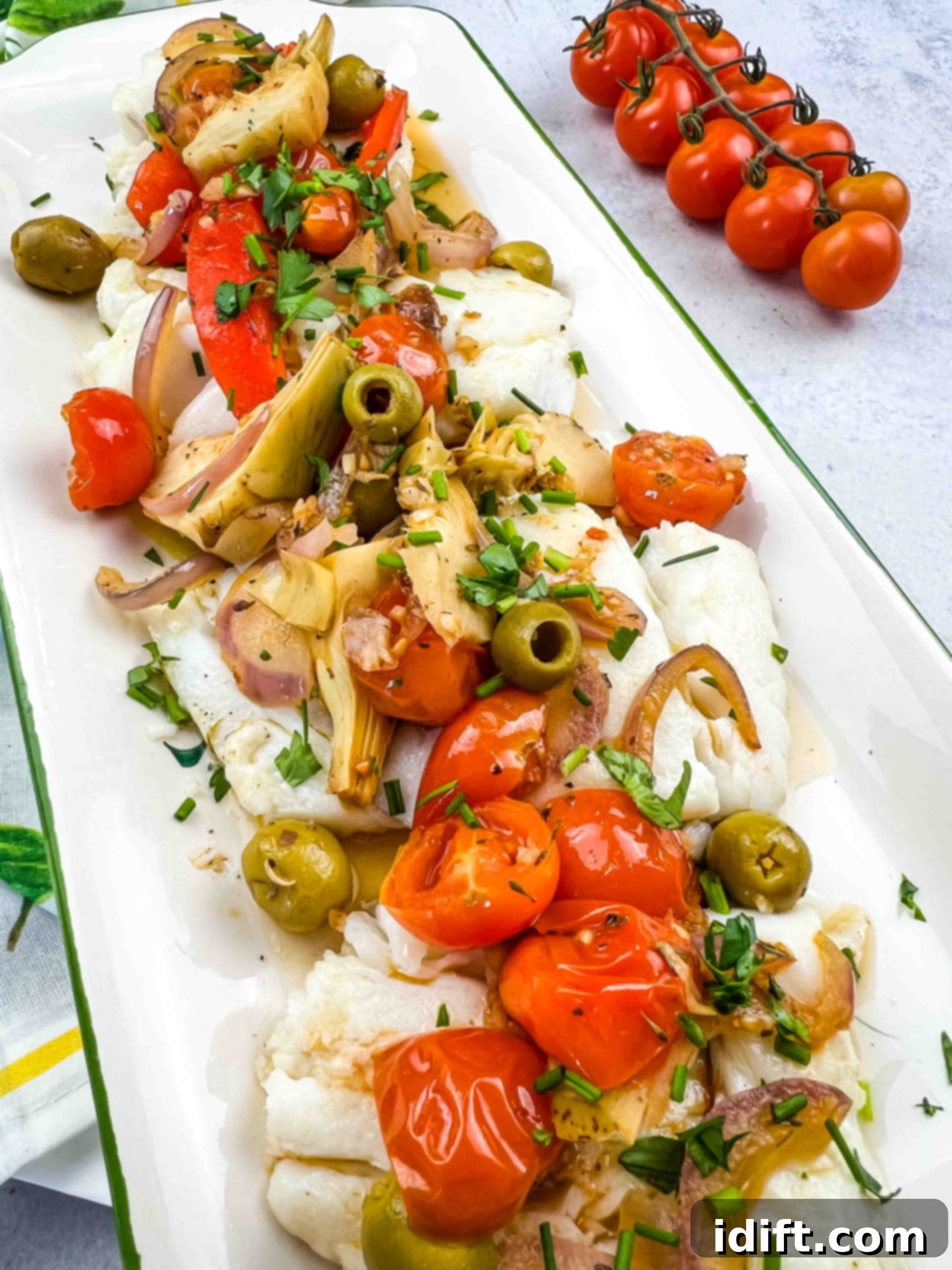 A beautifully plated Mediterranean Cod dish, showcasing flaky white fish surrounded by colorful cherry tomatoes, artichoke hearts, green olives, and fresh herbs.