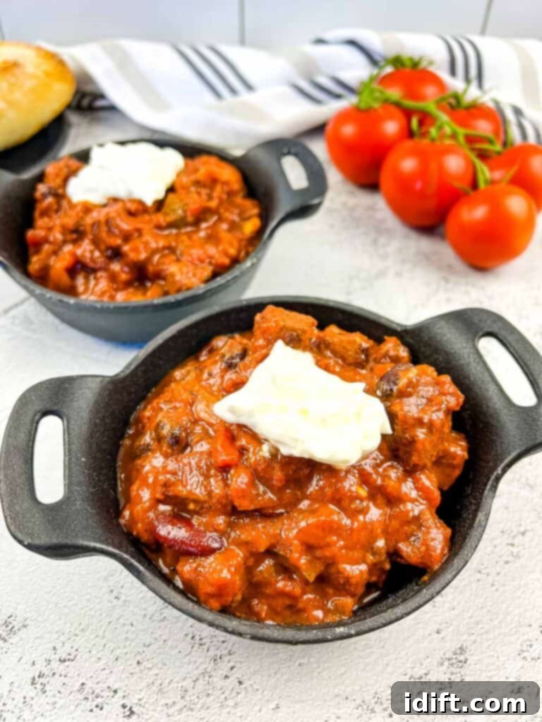 A closeup shot of Smoked Brisket Chili in 2 black bowls with bread and tomatoes in the background.