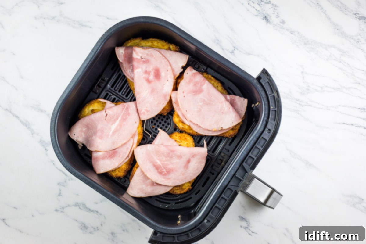 Partially cooked chicken cutlets in the air fryer basket, now topped generously with slices of deli ham.
