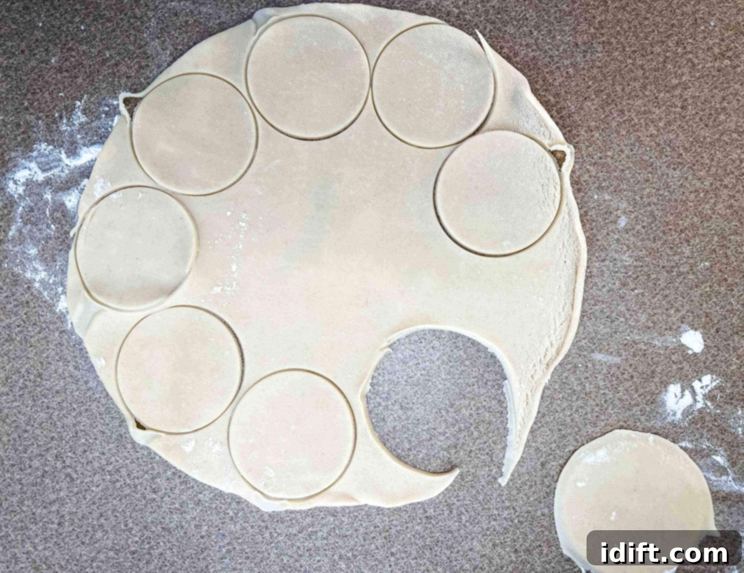 A person using a round cookie cutter to cut circular discs from a sheet of pie dough on a lightly floured surface.