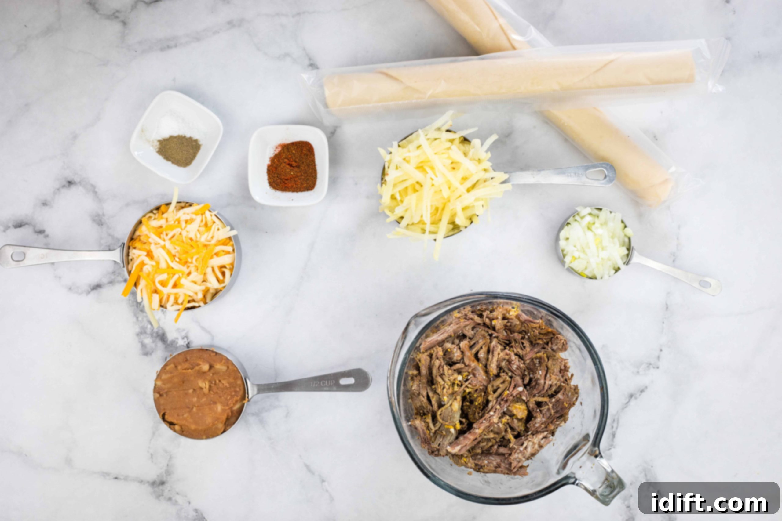 A spread of fresh ingredients laid out on a table, including bowls of shredded beef, cheese, potatoes, onions, refried beans, seasonings, pie dough, and an egg, ready for making air fryer beef empanadas.