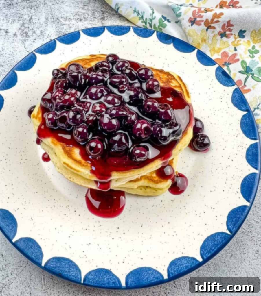 A tall stack of golden brown Blackstone Buttermilk pancakes, generously drizzled with vibrant blueberry syrup and topped with a pat of melting butter, served on a white plate.