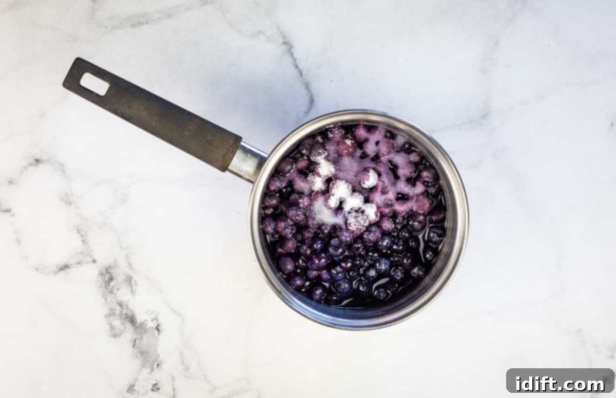 Blueberry sauce simmering in a small saucepan, with a whisk nearby, indicating the thickening process with cornstarch and water.