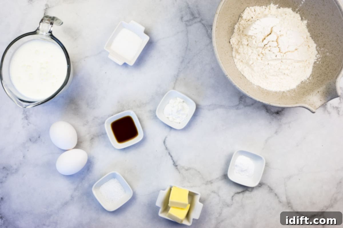 Various pancake ingredients laid out on a kitchen counter, including flour, baking powder, baking soda, salt, sugar, buttermilk, eggs, melted butter, and vanilla extract.