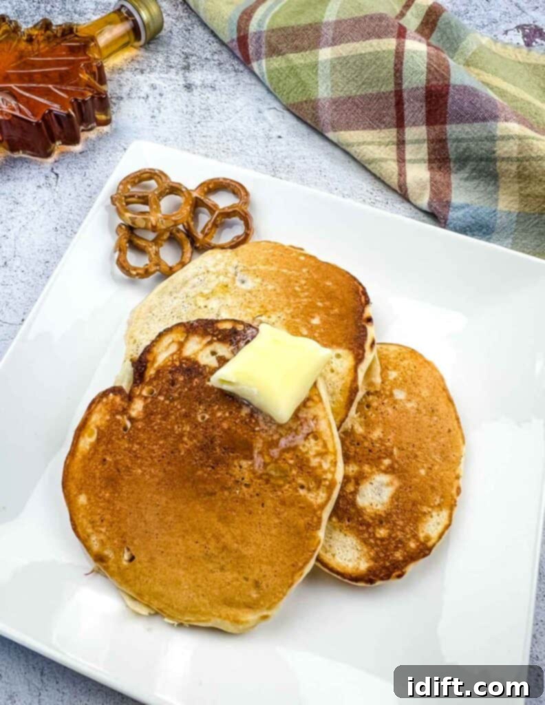 Golden brown pretzel pancakes stacked on a plate, adorned with a pat of melting butter and a drizzle of syrup, with more syrup visible in the background.