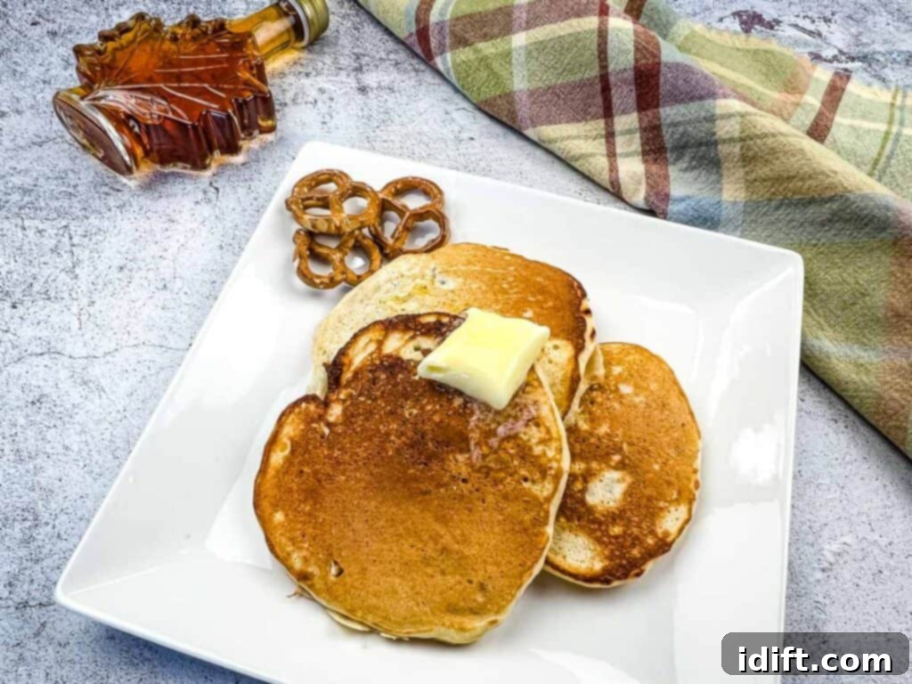 A horizontal shot of Pretzel Pancakes on a plate with melted butter and syrup drizzled over them. A syrup pitcher and fresh fruit are in the blurred background.