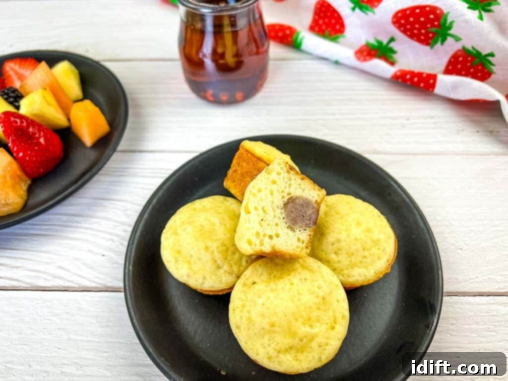 Sausage Pancake Poppers 3 Pancake bites with sausage artfully arranged on a black plate with fresh fruit and maple syrup in the background.