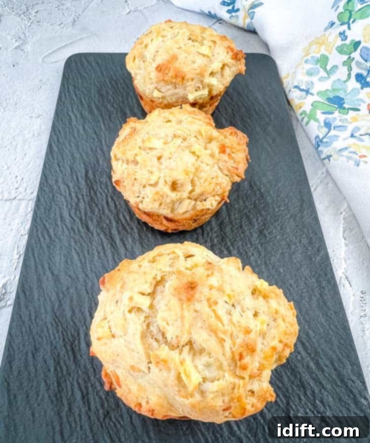 Three golden-brown Apple Cheddar Muffins, perfectly baked and inviting, displayed on a sleek black plate.