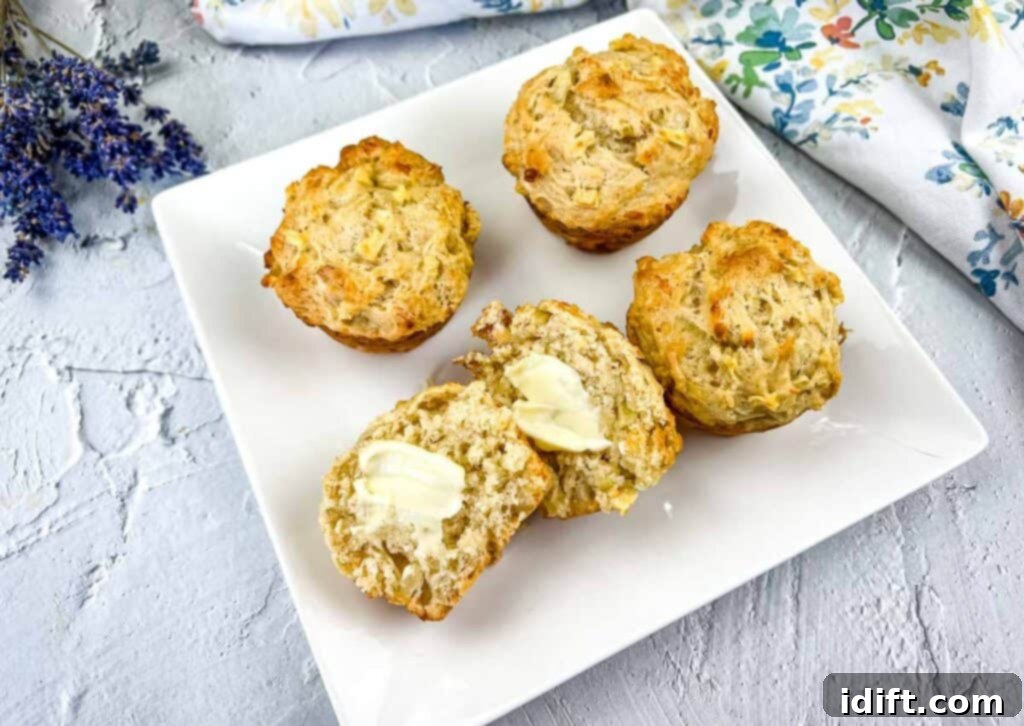 A close-up shot of an Apple Cheddar Muffin sliced in half, revealing its moist interior and apple chunks, served on a pristine white plate with a pat of melting butter.