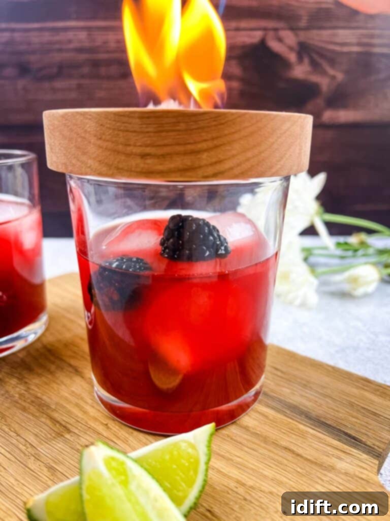 A cocktail smoker plate rests on top of a glass, with wood shavings lit by a torch, producing a plume of smoke.