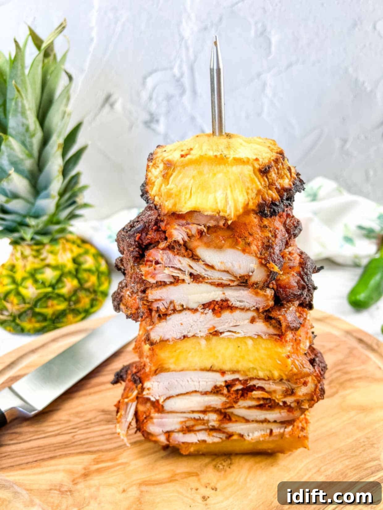 Smoked Chicken Al Pastor on a vertical skewer with pineapple in the background.