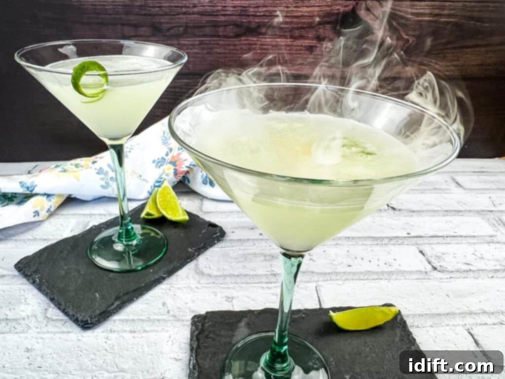 A horizontal shot of two elegant Smoked Gimlets, one in the foreground with wisps of smoke, and another blurred in the background.