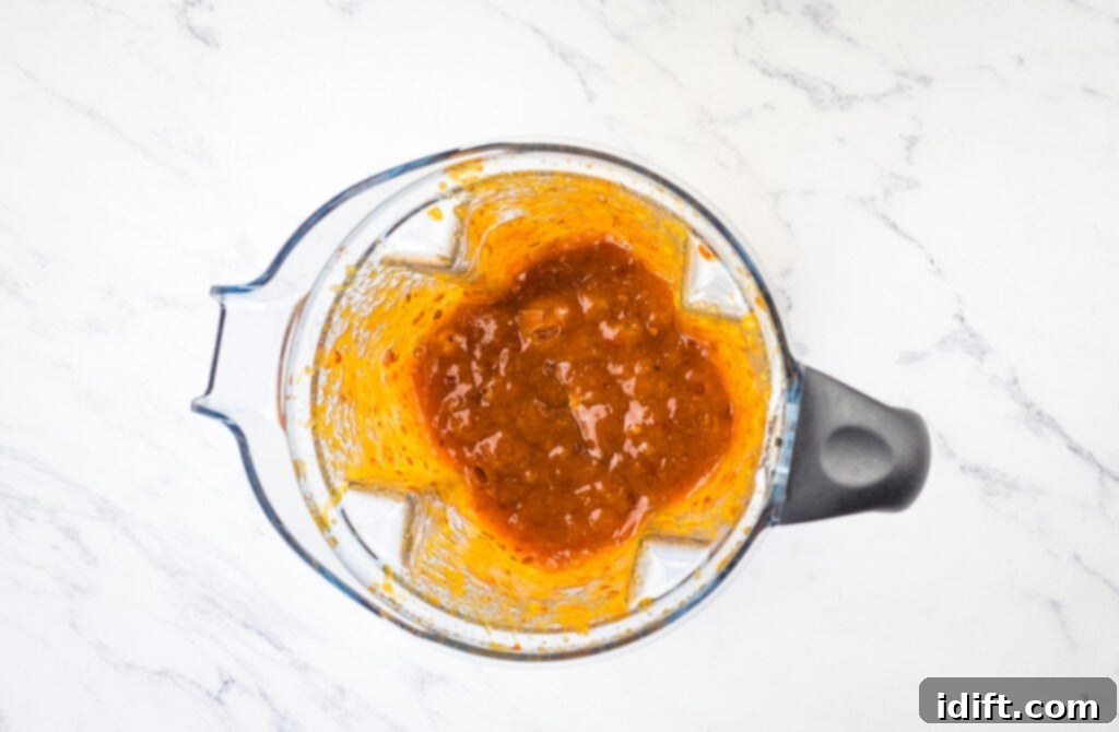 A blender filled with peach jam, chipotle, adobo sauce, and vinegar on a marble countertop, ready to be blended into a glaze.