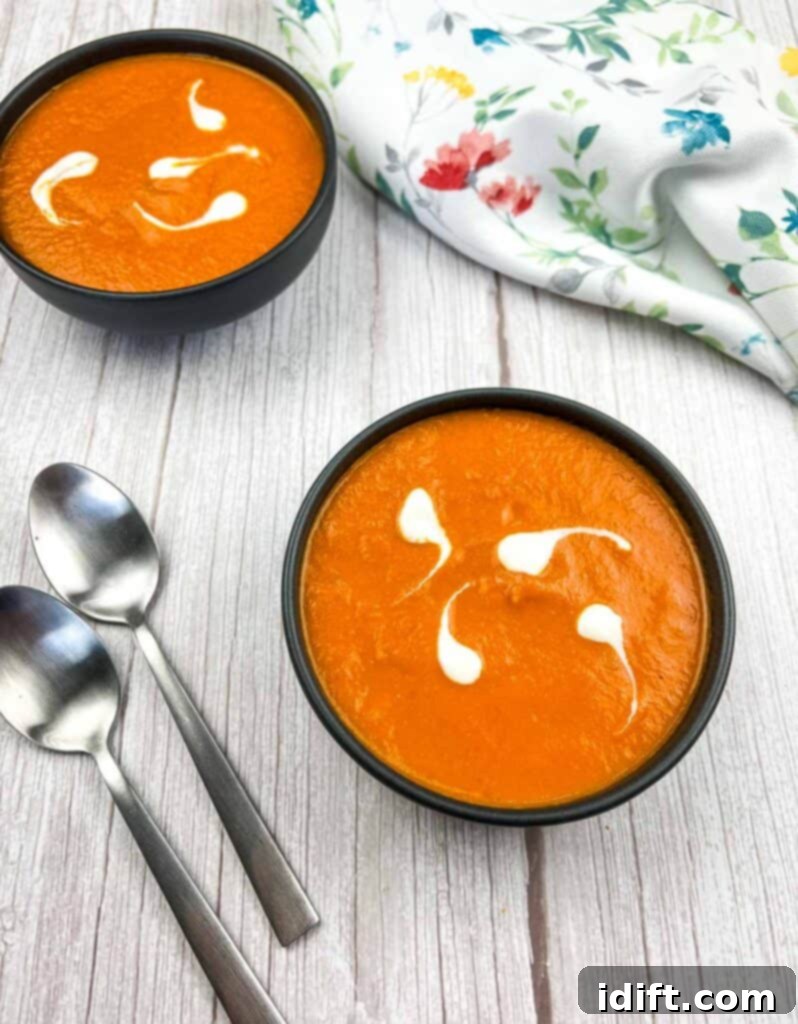 Two bowls of creamy oven-roasted tomato soup, garnished with fresh basil, on a rustic wooden table.