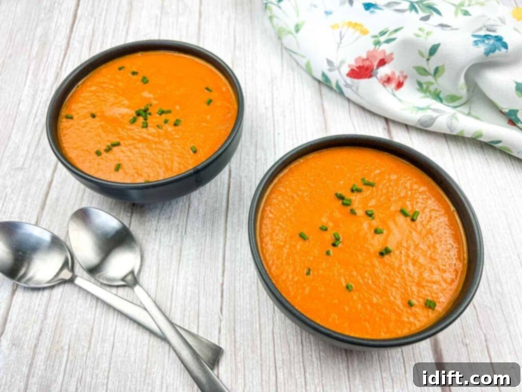 Two bowls of creamy oven-roasted tomato soup with spoons on a wooden table, garnished with herbs.