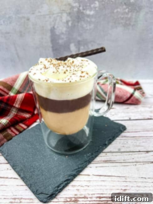 A stunning glass of Canadian Coffee, elegantly layered with rich coffee at the bottom, a creamy maple liqueur middle, and crowned with a generous swirl of whipped cream and delicate chocolate shavings. A cinnamon stick adds a touch of aromatic garnish.