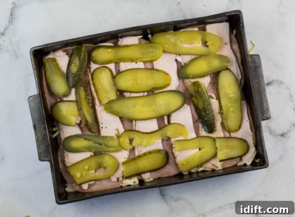 Cubano Casserole 12 Sliced dill pickles are spread over layers of ham and other ingredients in a baking pan, adding a crucial tangy element to the casserole.