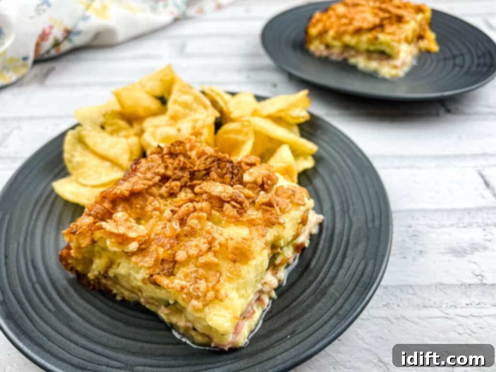 Cubano Casserole 4 A single, generous portion of Cuban Sandwich Casserole, featuring a golden-brown top and visible layers of meat and cheese, presented on a white plate with a side of crispy potato chips.