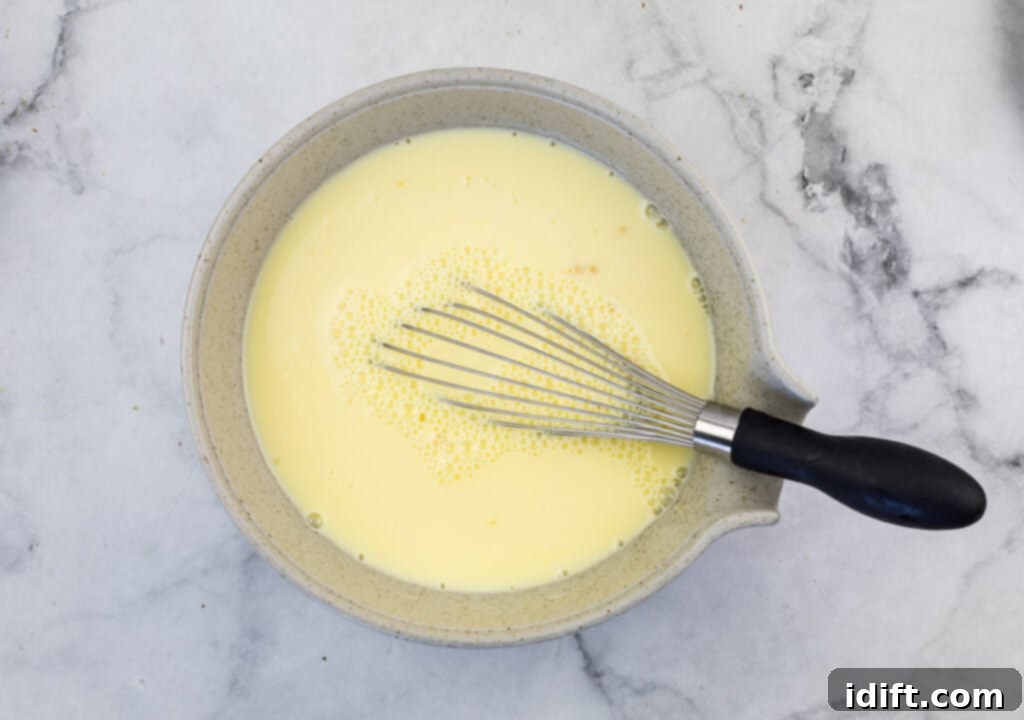 Cubano Casserole 7 A clear glass bowl containing a vibrant yellow mixture of egg yolks and milk, with a whisk resting inside, indicating the wet ingredients are being prepared.