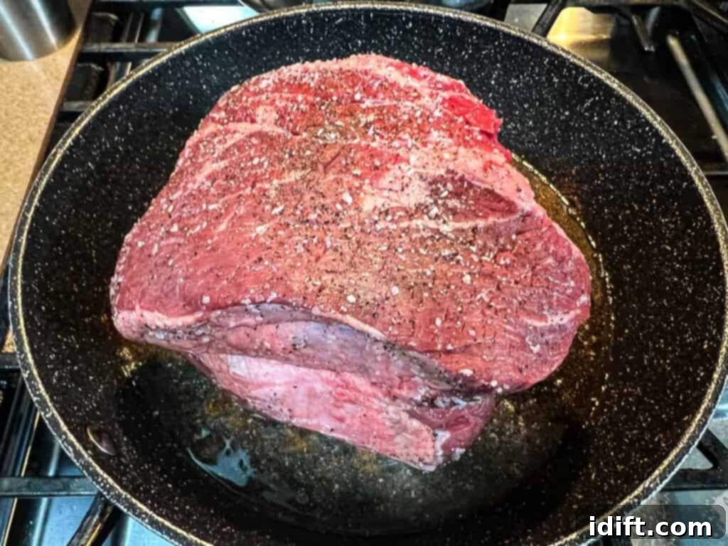 A piece of chuck roast being seasoned generously with salt and pepper in a frying pan before searing.