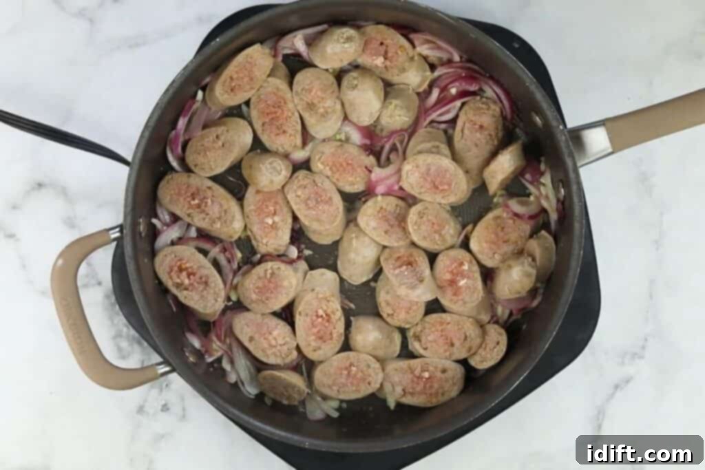 Sliced, par-cooked sausage browning in a skillet with sautéed onions and garlic.