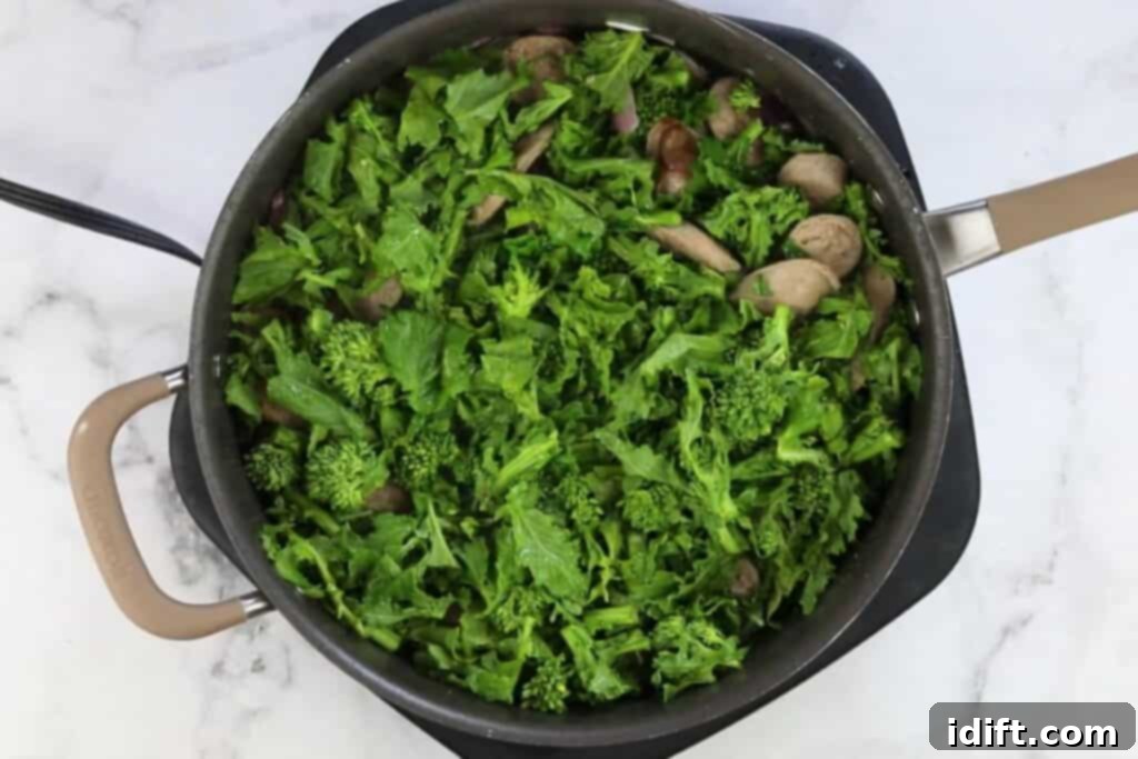 Freshly chopped broccoli rabe and chicken broth added to a skillet with sausage and onions, about to be simmered.