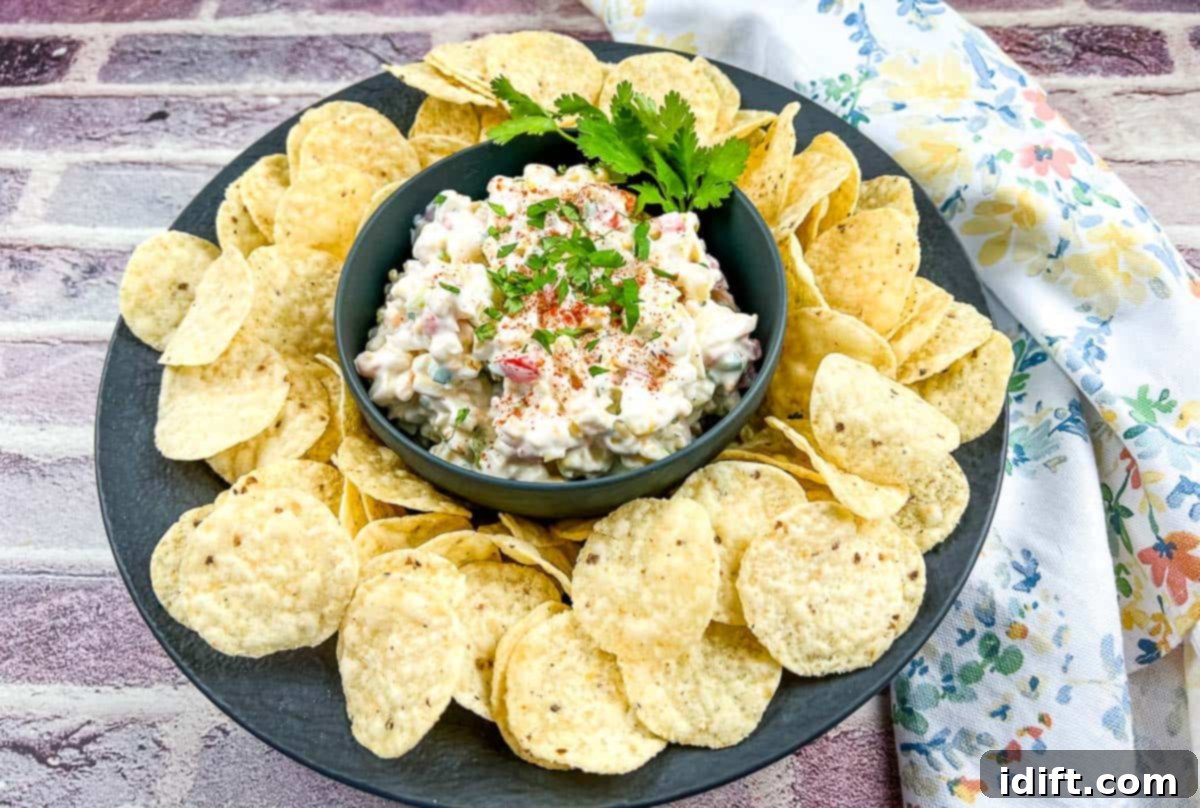 A rustic black plate featuring a bowl of refreshing Cold Corn Dip, surrounded by crunchy tortilla chips.