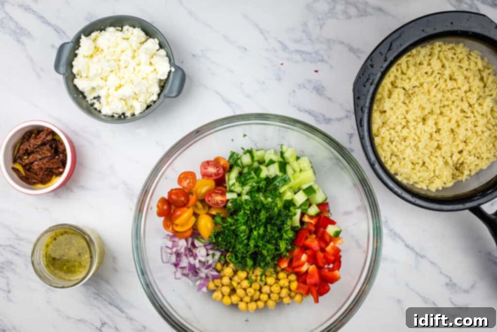 A large glass bowl filled with a colorful medley of chickpeas, grape tomatoes, and diced cucumbers, representing the initial vegetable prep for the Mediterranean Orzo Salad.