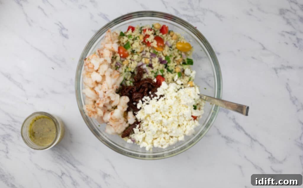 A large glass bowl containing a vibrant mix of cooked orzo pasta, fresh vegetables, tender shrimp, and crumbled feta cheese, awaiting the dressing.
