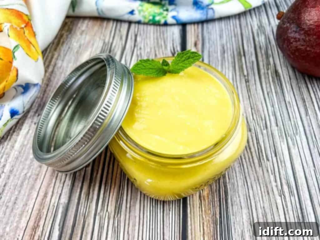 A rustic glass jar filled with vibrant, golden Mango Curd, accompanied by a fresh green mango and a sprig of mint.