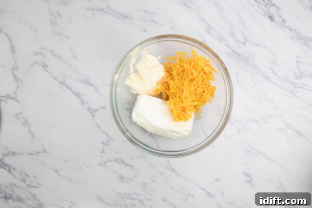 A glass bowl containing cream cheese, mayonnaise, and shredded cheddar, mixed together.