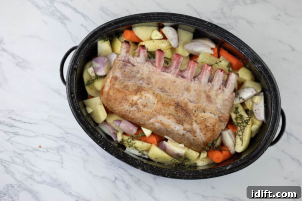 A roasting pan filled with colorful root vegetables and fresh thyme sprigs, with a perfectly seared pork roast nestled in the center, surrounded by a flavorful liquid base.